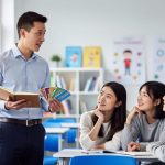 A Canadian English teacher stands in a bright classroom helping Chinese students learn with an open notebook and flashcards.